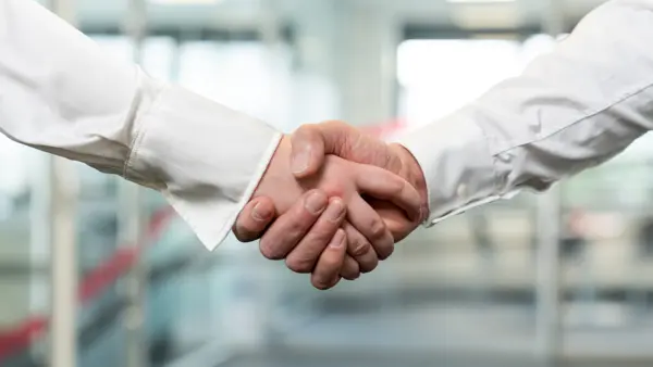 Zwei Personen in weißen Hemden geben sich die Hand vor einem unscharfen Bürohintergrund. Der Handschlag symbolisiert Partnerschaft, Zusammenarbeit und Vertrauen.